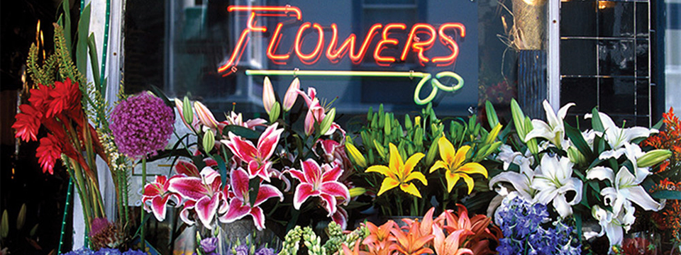 Photo of the front of a Flower Shop with the word 'Flowers' in neon in the window lit up just above rows and rows of flower arrangements.